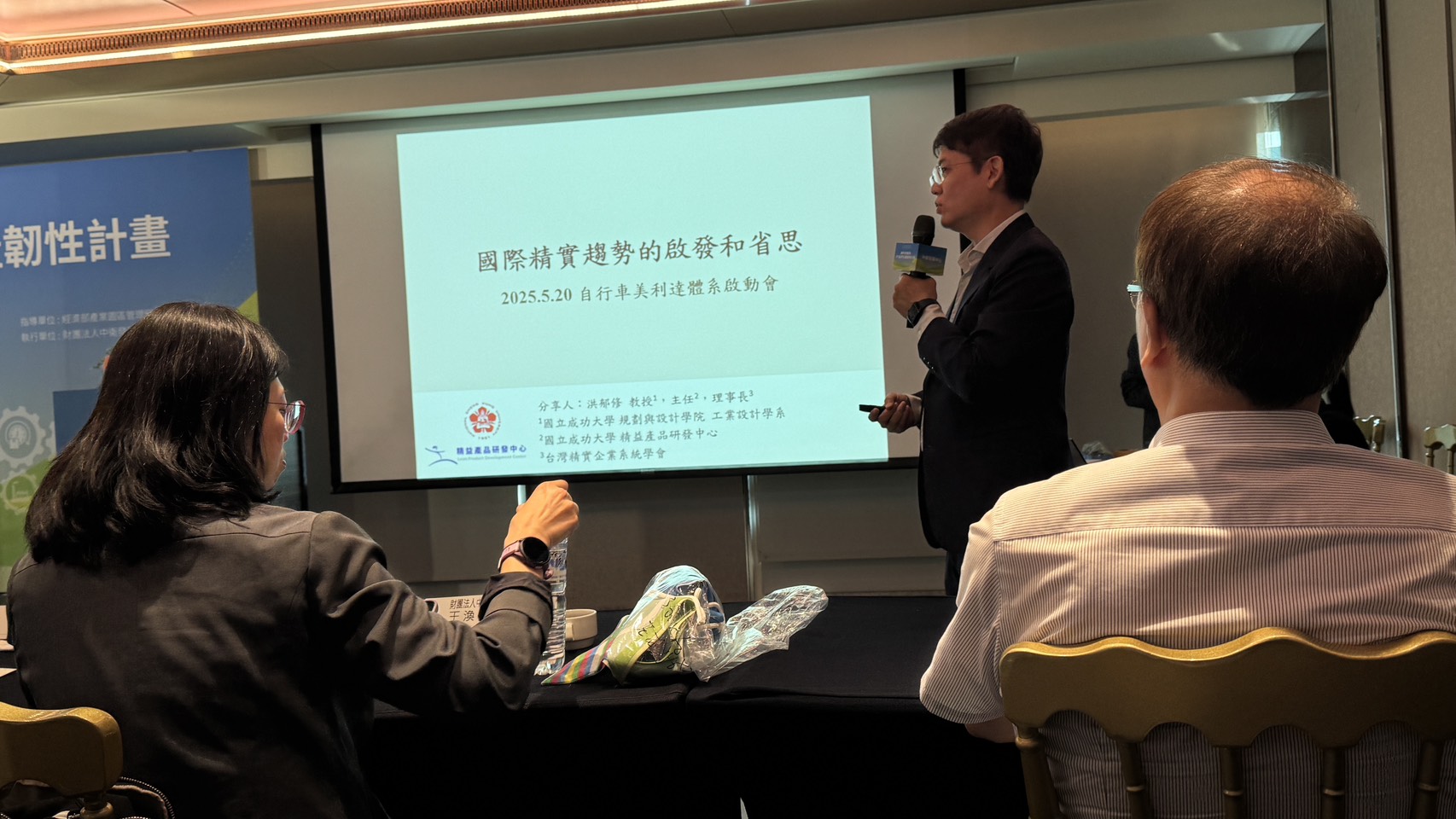 自行車美利達體系啟動會現場，洪郁修教授在台前分享，投影片標題為「國際精實趨勢的啟發和省思」，左側背板為經濟部產業發展署指導、中衛發展中心執行的韌性計畫